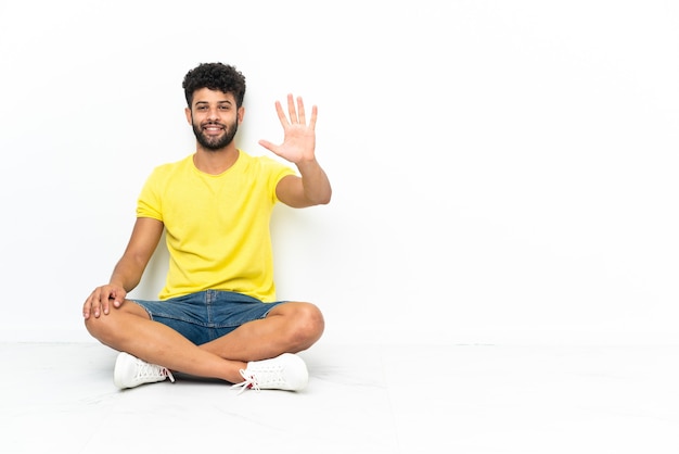 Młody przystojny mężczyzna marokański siedzi na podłodze na białym tle, licząc pięć palcami