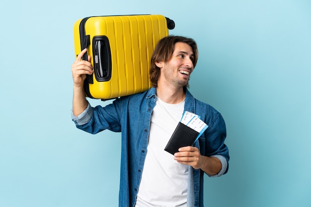 Młody przystojny mężczyzna na białym tle na niebieskiej ścianie w wakacje z walizką i paszportem