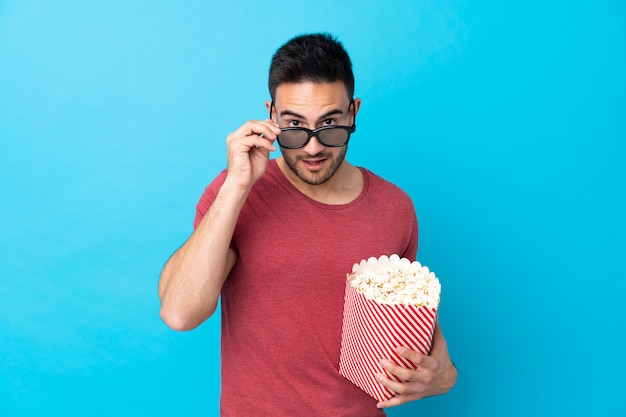 Młody przystojny mężczyzna na niebiesko zaskoczony okularami 3d i gospodarstwa duże wiadro popcornów