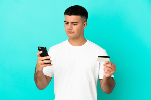 Młody przystojny mężczyzna nad odosobnioną niebieską ścianą kupując telefon komórkowy za pomocą karty kredytowej