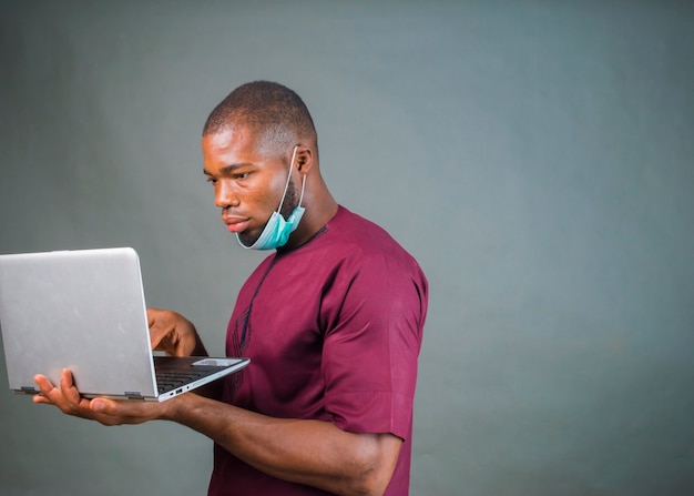 Młody przystojny mężczyzna nigeryjski na białym tle nad szarym tle noszenie maski i za pomocą swojego laptopa z powodu wybuchu wirusa.