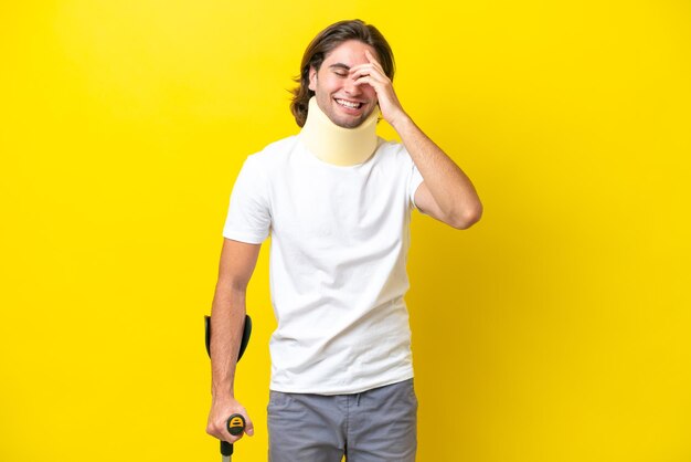 Młody przystojny mężczyzna noszący ortezę na szyję i kule odizolowany na żółtym tle śmiejąc się