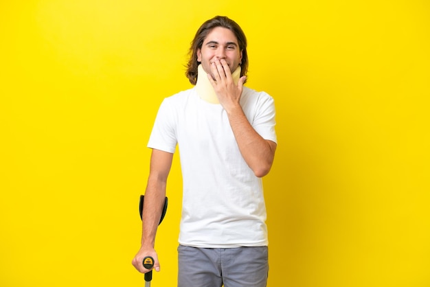 Młody przystojny mężczyzna noszący ortezę na szyję i kule odizolowany na żółtym tle szczęśliwy i uśmiechnięty zakrywający usta dłonią