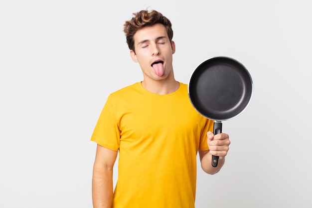 Młody przystojny mężczyzna o radosnym i buntowniczym nastawieniu, żartujący, wystawiający język i trzymający patelnię kucharską
