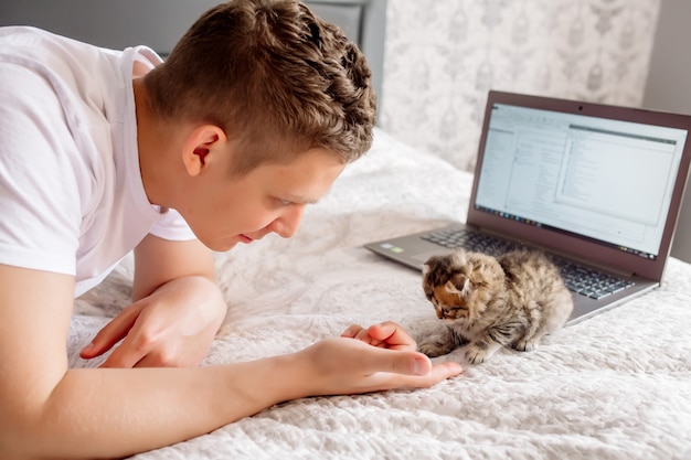 Zdjęcie młody przystojny mężczyzna pracujący w domu podczas kwarantanny koronowirusa, leżący z laptopem i kotkiem w łóżku