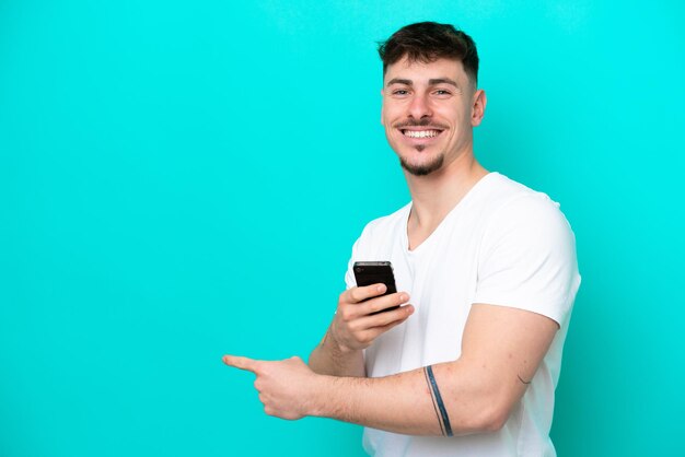 Młody przystojny mężczyzna rasy kaukaskiej na białym tle na niebieskim tle za pomocą telefonu komórkowego i wskazując wstecz