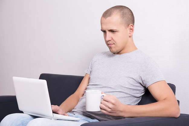 Młody przystojny mężczyzna siedzi na kanapie z komputerem i filiżanką herbaty lub kawy