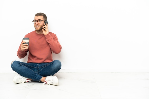 Młody przystojny mężczyzna siedzi na podłodze trzymając kawę na wynos i telefon komórkowy