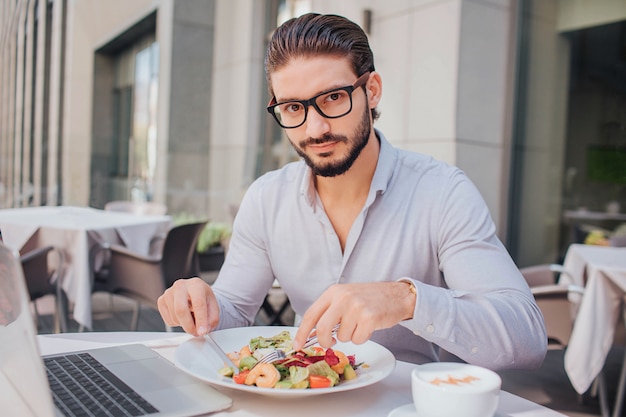 Młody przystojny mężczyzna siedzi przy stole i je lunch. Patrzy w kamerę. Facet pozuje. On jest gotowy do jedzenia sałatki. Przy stole jest laptop i filiżanka kawy.