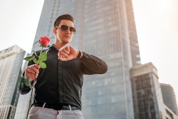 Młody przystojny mężczyzna stoi przed drapaczem chmur i patrzy na zegarki. W ręku trzyma czerwoną różę.