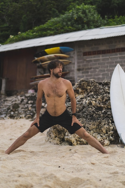 Młody przystojny mężczyzna surfer na brzegu oceanu robi rozgrzewkę przed surfowaniem. ćwiczenia przed sportem, stretching przed surfingiem.