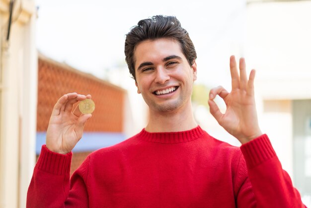 Młody przystojny mężczyzna trzyma Bitcoina na zewnątrz, pokazując znak ok palcami
