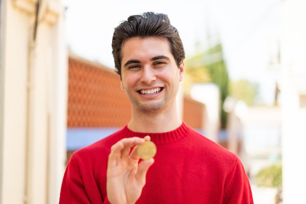 Młody przystojny mężczyzna trzyma Bitcoina na zewnątrz z radosnym wyrazem twarzy