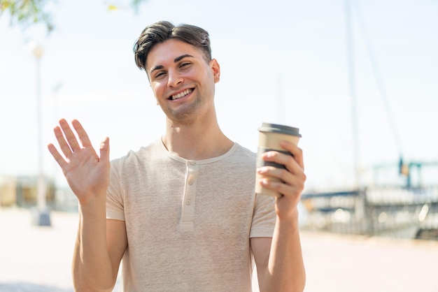 Młody przystojny mężczyzna trzyma kawę na wynos na zewnątrz pozdrawiając ręką z radosnym wyrazem twarzy