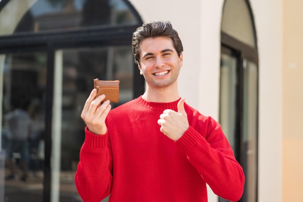 Młody przystojny mężczyzna trzyma portfel na zewnątrz z kciukami do góry, ponieważ stało się coś dobrego