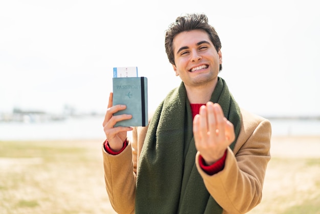 Młody przystojny mężczyzna trzymający paszport na zewnątrz zapraszający do przyjścia z ręką Szczęśliwy, że przyszedłeś