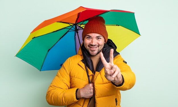 Młody przystojny mężczyzna uśmiecha się i wygląda przyjaźnie, pokazując numer dwa. koncepcja deszczu i parasola