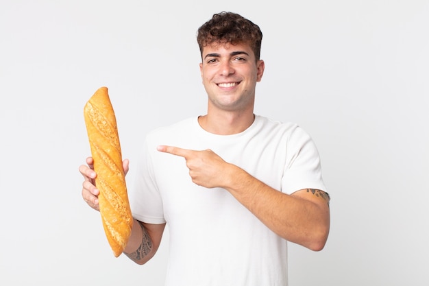 Młody przystojny mężczyzna uśmiecha się radośnie, czuje się szczęśliwy, wskazuje na bok i trzyma bagietkę z chlebem