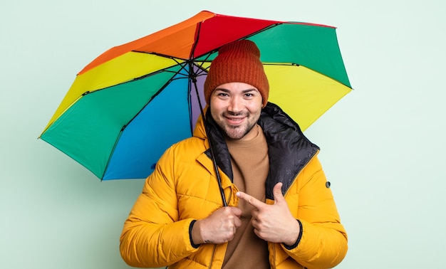 Młody przystojny mężczyzna uśmiechający się radośnie, czując się szczęśliwy i wskazując na bok. koncepcja deszczu i parasola