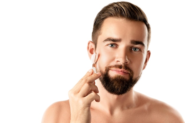 Młody przystojny mężczyzna używa wałka do twarzy do masażu twarzy na białym tle