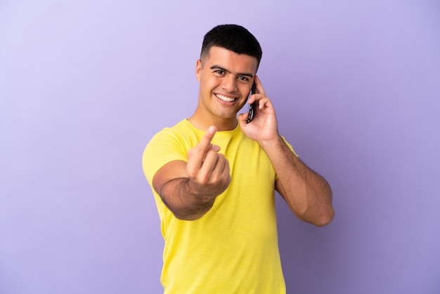 Młody przystojny mężczyzna używający telefonu komórkowego na odosobnionym fioletowym tle robi nadchodzący gest