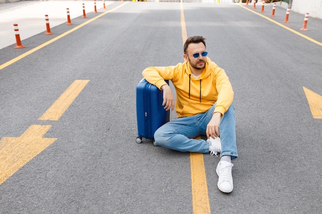 młody przystojny mężczyzna w żółtym ubraniu z niebieską walizką na drodze