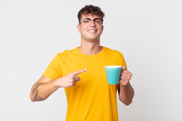Młody przystojny mężczyzna wyglądający na podekscytowanego i zdziwionego, wskazując na bok i trzymający filiżankę kawy