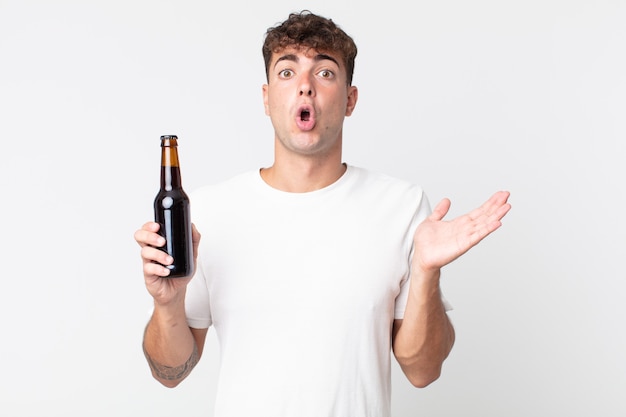 Młody przystojny mężczyzna wyglądający na zaskoczonego i zszokowanego, z opuszczoną szczęką, trzymający przedmiot i trzymający butelkę piwa