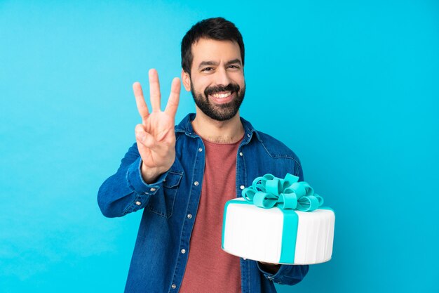Młody przystojny mężczyzna z dużym tortem nad odosobnioną błękit ścianą szczęśliwą i liczy trzy z palcami