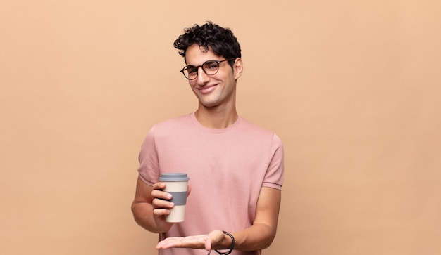 Młody przystojny mężczyzna z kawą uśmiechający się radośnie, czujący się szczęśliwy i pokazujący koncepcję w przestrzeni kopii z dłonią