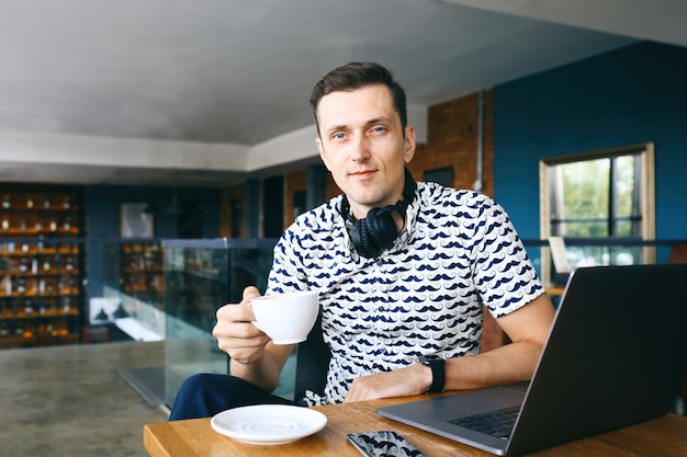 Młody przystojny modnisia mężczyzna siedzi w kawiarni, trzyma filiżankę kawy. Laptop i telefon komórkowy na drewnianym stole.