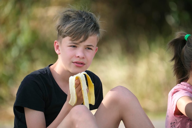 Młody przystojny nastolatek chłopiec je smacznego dojrzałego banana podjadającego na zewnątrz w letni dzień