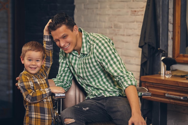 Młody przystojny ojciec i jego mały stylowy syn u fryzjera czekając na fryzjera. Ojciec siedzi na krześle, a obok stoi syn