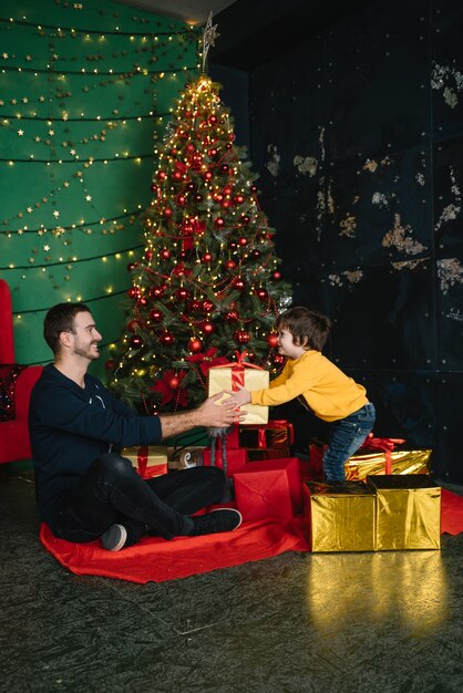 Młody przystojny ojciec z uroczym synem w pobliżu choinki z prezentami. Wesołych Świąt