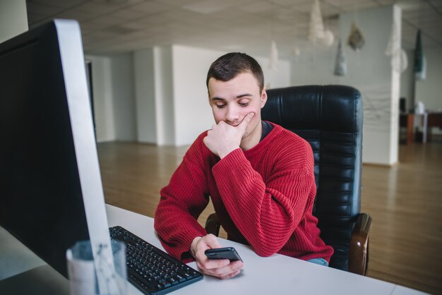 Młody przystojny projektant hipster siedzi przy monitorze komputera i użyj telefonu komórkowego. Życie biurowe.