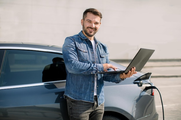 Młody Przystojny Uśmiechnięty Mężczyzna Pracuje Na Laptopie, Czekając, Aż Jego Luksusowy Samochód Elektryczny ładuje Się, Płacąc Za ładowanie Stojąc W Pobliżu Stacji Miejskiej Z Kablem Zasilającym Podłączonym Do Samochodu