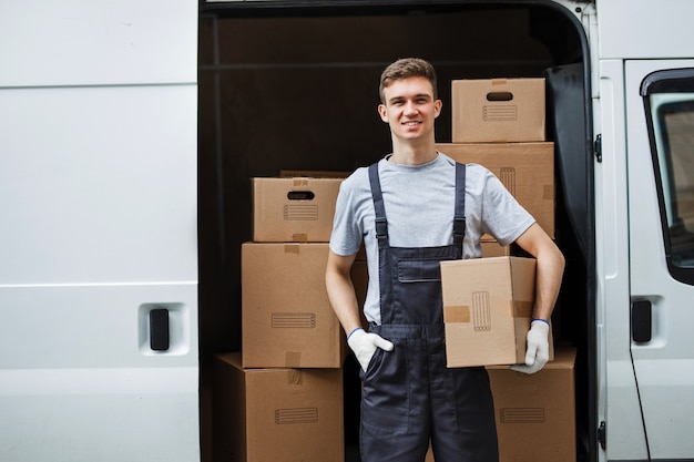 Zdjęcie młody przystojny uśmiechnięty pracownik w mundurze stoi obok furgonetki pełnej pudeł, trzymając w rękach pudełko. przeprowadzki, usługi przewozowe.