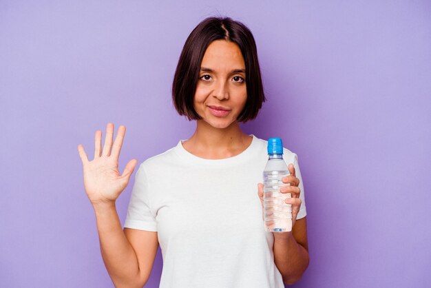 Młody rasy mieszanej trzymając butelkę wody na białym tle na fioletowym tle uśmiechnięty wesoły pokazując numer pięć palcami.