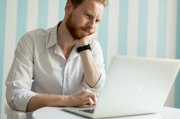 Młody redhair mężczyzna pracuje na laptopie w pokoju błękitną pasiastą ścianą