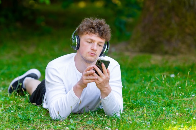 Zdjęcie młody rudowłosy mężczyzna w słuchawkach leży na zielonej trawie patrząc na telefon