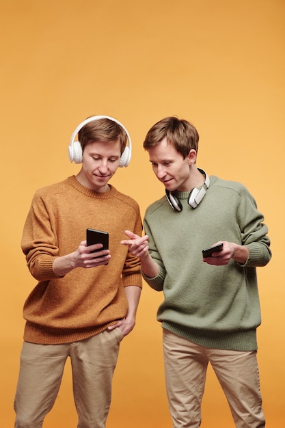 Młody rudy mężczyzna w bezprzewodowych słuchawkach pokazujący ekran telefonu i udostępniający utwór muzyczny z bratem bliźniakiem