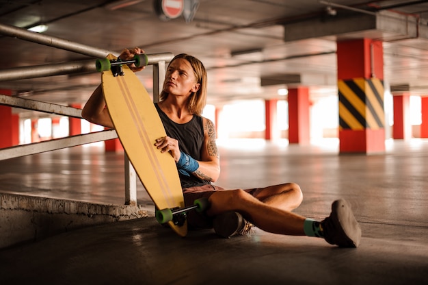 Młody rudzielec mężczyzna siedzi na podłoga parking i trzyma longboard