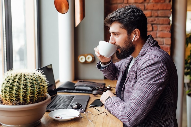 Młody Skoncentrowany Brodaty Biznesmen W Słuchawkach, Używający Laptopa I Pijący Kawę Latte