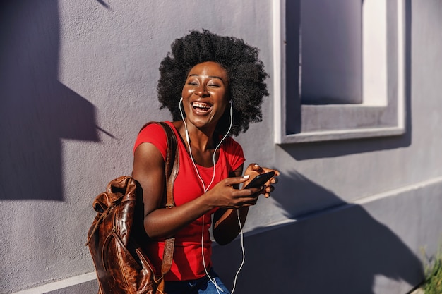 Młody śmiejąc się afrykańskiej kobiety stojącej na zewnątrz, trzymając inteligentny telefon i słuchając muzyki