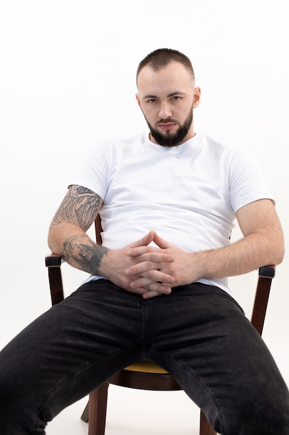 Młody spokojny brodaty mężczyzna z krótkimi ciemnymi włosami w białej koszulce, czarnych dżinsach siedzi na drewnianym krześle inte