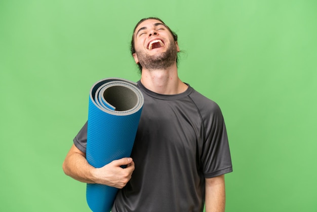 Zdjęcie młody sportowiec idzie na zajęcia jogi, trzymając matę na odizolowanym tle śmiejąc się
