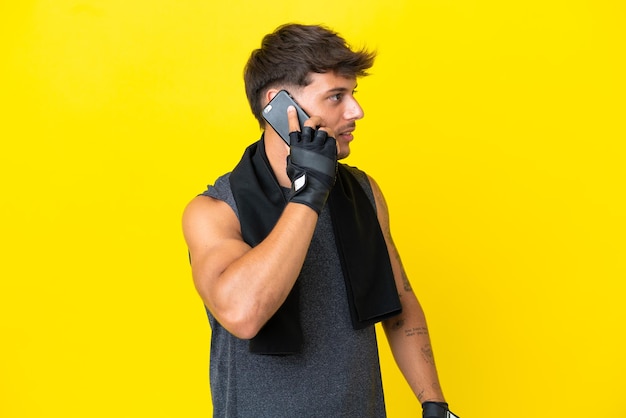 Młody sportowiec kaukaski z ręcznikiem na białym tle na żółtym tle, który prowadzi rozmowę z telefonem komórkowym z kimś