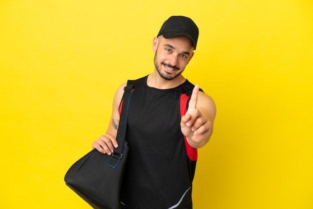 Młody sportowiec kaukaski z torbą sportową na białym tle na żółtym tle pokazujący i unoszący palec
