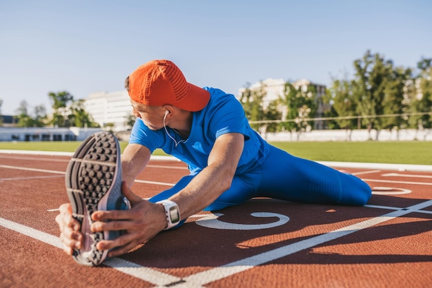 Młody sportowiec mężczyzna rozciągając nogę na torze na stadionie przygotowuje się do treningu biegania i joggingu Kaukaski mężczyzna ćwiczenia na zewnątrz na sobie niebieską odzież sportową Styl życia ludzi sportu