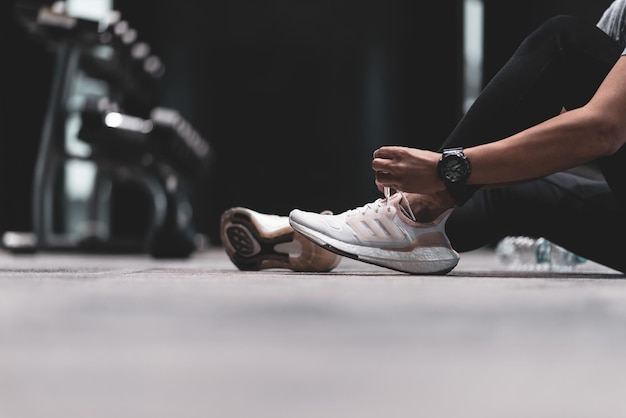 Zdjęcie młody sportowiec w czarnych spodniach do ćwiczeń siedzi na podłodze w siłowni, wiąże swoje białe buty do biegania w przygotowaniu do porannego biegania.